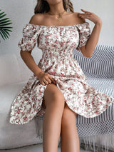 Donna seduta con abito estivo floreale bianco e rosa, spalle scoperte, in un ambiente accogliente. Moda femminile, stile casual elegante.