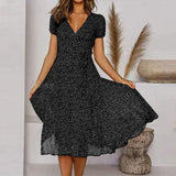 Abito nero a fiori con scollo a V e maniche corte, indossato da una donna in un ambiente moderno con decorazioni naturali. Moda femminile elegante.