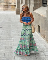 Donna sorridente con abito lungo estivo a fantasia geometrica e floreale, occhiali da sole bianchi, borsa di paglia, cammina su strada cittadina.