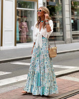 Donna con camicia bianca annodata e gonna lunga floreale azzurra, occhiali da sole bianchi, borsa di paglia, cammina in città. Moda estiva elegante.