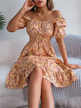 Donna seduta su divano indossa abito estivo floreale giallo con spalle scoperte, maniche a sbuffo e gonna arricciata. Moda femminile elegante.