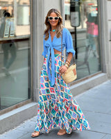 Donna con camicia azzurra annodata, gonna lunga colorata, occhiali da sole bianchi e borsa di paglia. Moda estiva urbana, look casual chic.